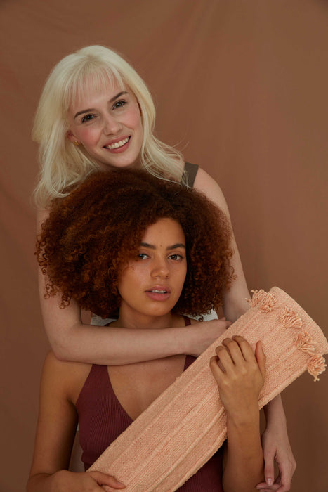 Two women standing close together against a brown background, one holding a rolled up yoga mat.