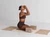 Woman in brown athletic wear practicing yoga on a mat with yoga blocks against a plain background