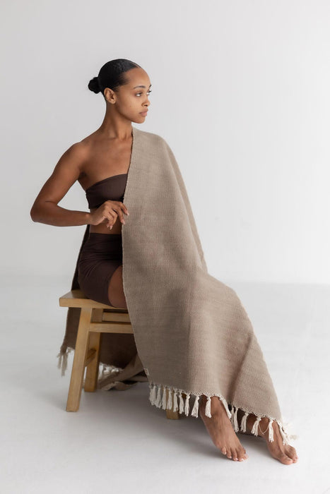 Woman sitting on a stool wearing a brown strapless top and draped in a beige yoga mat with tassels.
