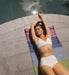 Woman in a white bikini lying on a colorful yoga mat by a pool.