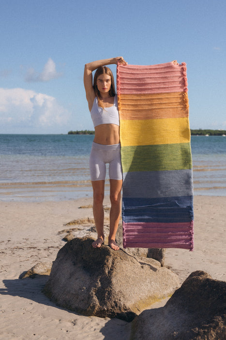 Woman holding up a colorful yoga mat on a beach with ocean and sky in the background