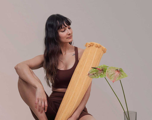 Woman holding a rolled up yoga mat with a plant in the foreground against a plain background