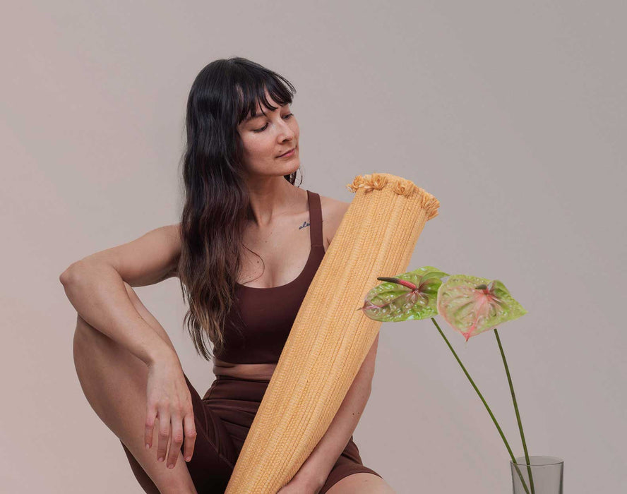 Woman holding a rolled up yoga mat with a plant in the foreground against a plain background