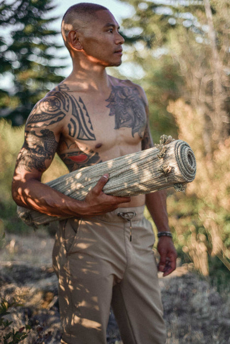 Man with tattoos holding a rolled up yoga mat in a forest setting